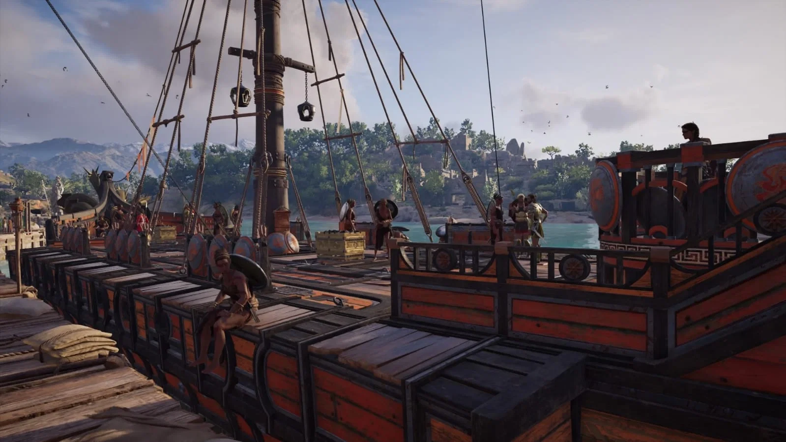 The image depicts a highly detailed scene of an ancient Mediterranean harbor, viewed from the dock alongside a large wooden ship that occupies most of the foreground. The vessel appears to be either a warship or a heavily armed transport, constructed from dark timber with striking red-orange painted panels bordered by black wooden frames. Its design reflects classical Greek naval architecture, with decorative round shields mounted along the sides of the deck. These shields feature weathered blue-gray surfaces accented by orange-red patterns, adding both practical and ornamental value to the ship's appearance. The deck is filled with neatly stacked wooden cargo crates of varying sizes, suggesting that the ship is carrying supplies, trade goods, or military provisions. Some larger chests reinforced with metal bands are positioned near the center of the deck, while smaller crates line the outer edges in organized rows. The arrangement creates the impression of a vessel preparing for a voyage or having recently unloaded part of its cargo. Rising from the center of the ship is a tall, imposing mast surrounded by an intricate network of ropes, rigging, spars, pulleys, and hanging fittings. The complex web of lines stretches in multiple directions, supporting the ship's sails and giving the vessel an authentic, functional appearance. The rigging dominates the upper portion of the image, drawing the eye upward and emphasizing the ship's impressive scale. Several people are scattered across the deck, bringing the harbor scene to life. In the foreground, a shirtless crew member sits casually on one of the cargo crates, resting with one leg hanging over the side of the ship. His relaxed posture contrasts with the activity elsewhere on deck. Near the center, two individuals stand beside the larger cargo chests, appearing to converse or inspect the supplies. On the right side of the ship, a small group of four figures gathers near the railing. One of them wears noticeably more elaborate armor with golden accents, suggesting a person of higher rank, such as an officer or captain, while the others are dressed in simpler tunics and light armor typical of sailors or soldiers. At the far right, another individual stands on an elevated platform near the stern, gazing out over the harbor as if overseeing preparations or watching activity along the waterfront. To the left, the dock continues alongside the ship. Wooden planks form the pier, where several large sacks are stacked in the immediate foreground. Another similarly styled vessel is partially visible further down the harbor, while additional people can be seen moving about the docks, contributing to the atmosphere of a busy port. Beyond the ship lies calm, crystal-clear turquoise water reflecting the daylight. Across the harbor, a lush shoreline stretches into the distance, densely covered with green trees. Nestled among the vegetation are ancient stone buildings and ruins, their weathered walls partially hidden by the forest. In the far background, rugged mountains rise beneath the horizon, their bluish-gray slopes fading softly into the distance and adding a strong sense of depth to the landscape. Above, the sky is bright yet partly cloudy. Large, soft gray and white clouds drift across the scene while sunlight breaks through, casting warm highlights over the ship's wooden surfaces and illuminating the surrounding landscape. Numerous small birds circle high overhead, adding movement and realism to the otherwise peaceful setting. The image is rich in warm earth tones and natural colors. The dark wood of the ship contrasts beautifully with the vibrant red-orange decorative paint, the brilliant turquoise sea, the lush green vegetation, and the cool blue-gray mountains in the distance. The careful lighting and realistic textures create a cinematic atmosphere, making the harbor feel alive and immersive. Overall, the scene conveys the impression of an ancient maritime world at the height of its activity. The combination of detailed ship construction, organized cargo, interacting crew members, tranquil coastal scenery, and classical architecture creates a vivid snapshot of life in an ancient Mediterranean harbor, suggesting a moment just before departure or shortly after arrival during a trading or military expedition.